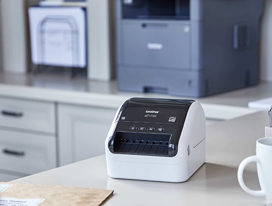 QL-1100C label printer sitting on a desk with Brother printer in background. 