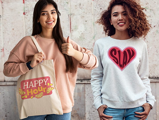 A Woman showcasing bag with imprinted "Happy Hour" design with another woman wearing a sweatshirt with a red heart and the word "Slay" written in it